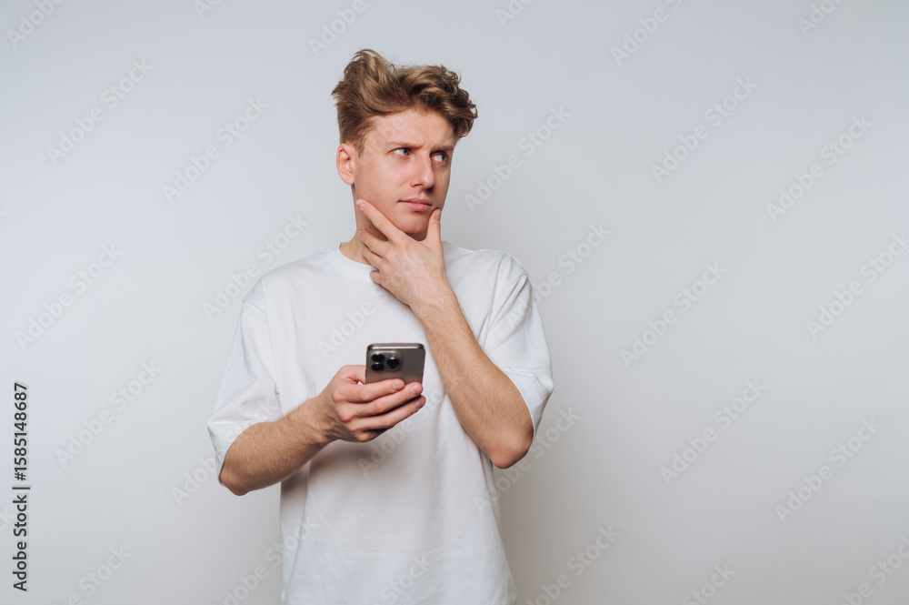 Young man contemplating while holding smartphone in a minimalistic studio environment Young man contemplating while holding smartphone in a minimalistic studio environment