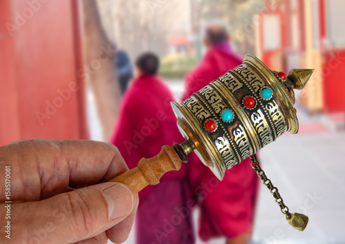 Buddhist Scripture Wheel, hand held spinning Prayer Wheel in front of two blurred Buddhist monks. The cylinder turn around a handle with a slight rotation of the wrist.