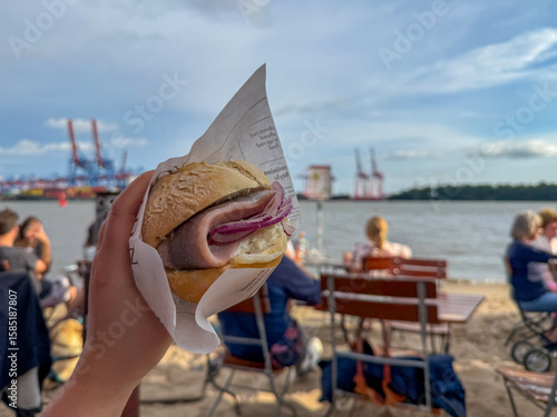 Traditional north german street food snack Fischbroetchen wheat bun with matjes fish fillet and onion in female hand close on Elbstrand beach