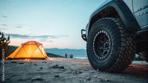 Off road adventure camping with illuminated tent and rugged vehicle tire at dusk
