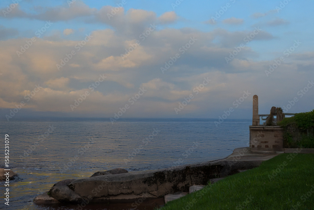 Fototapeta premium Calm sea view at sunset with dramatic clouds, a stone terrace, and lush grass in the foreground, creating a peaceful coastal atmosphere.