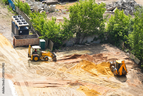 Two yellow excavators working on a construction site, moving sand and soil near trees and heavy equipment. Aerial top-down photo of groundwork, earthmoving and site preparation for development