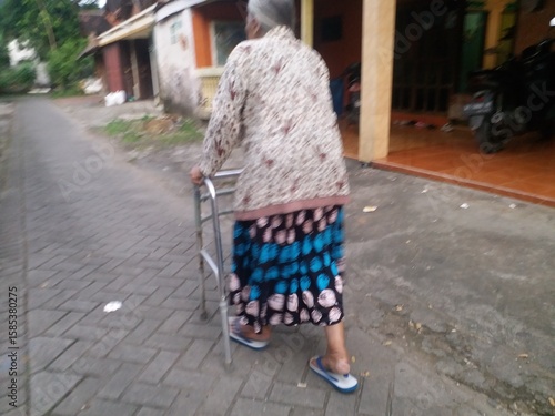 A senior woman wearing traditional Indonesian batik clothing is seen using a walker to assist her while walking on a sidewalk.
