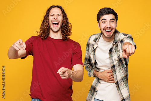 Young two friends buddies men they wearing casual clothes together pointing finger on camera with smile mocking or humiliate isolated on plain yellow orange color background studio. Lifestyle concept.