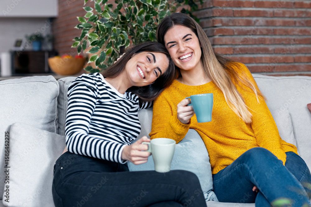 Obraz premium Two smiling young women talking while drinking coffee sitting on couch in the living room at home.