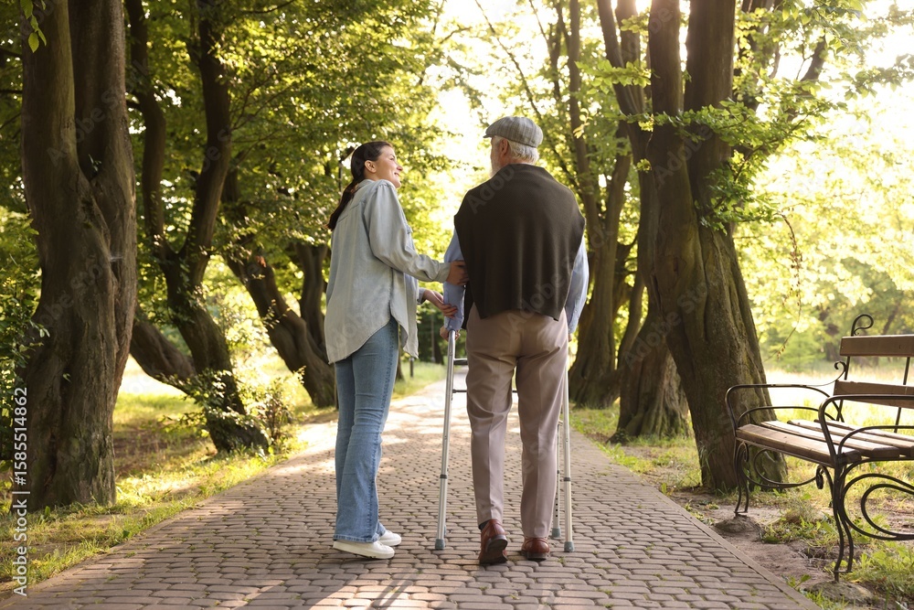 Fototapeta premium Smiling caregiver supporting elderly man who using walking frame outdoors, back view