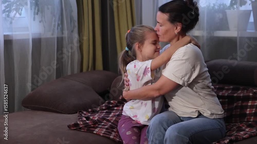 Mom hugs and kisses her eight-year-old little daughter while sitting on the couch at home in the evening. Concept of friendly family.