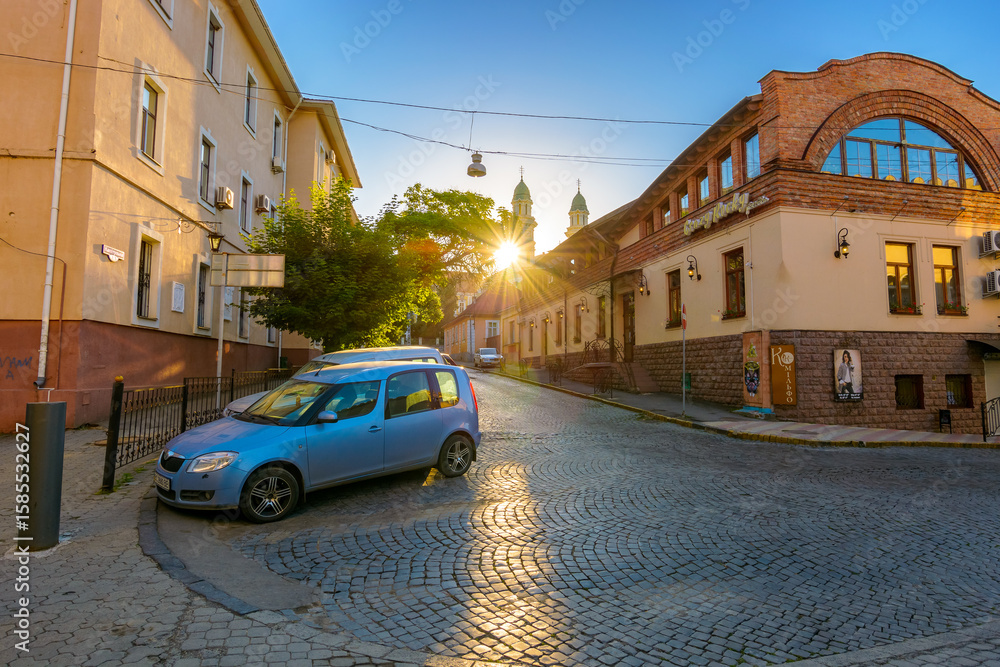 Obraz premium uzhhorod, ukraine - 04 jun 2017: european urban landscape with cars on street of town in morning light. sunrise above roof. holy cross cathedral in the distance. old cobblestone