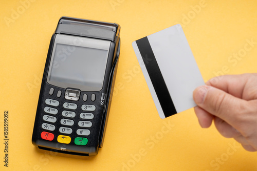 A person is holding a credit card in front of a payment terminal. The terminal has a green, yellow, and red button