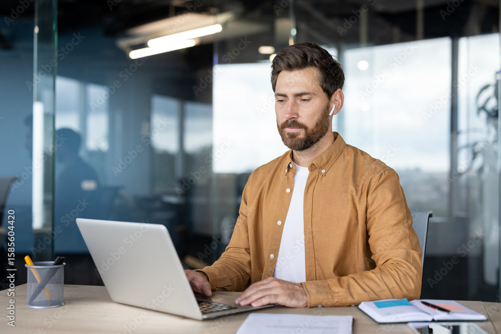 Fototapeta premium A focused man works on a laptop at a desk in a modern office environment, wearing earbuds.
