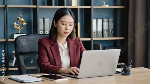 Asian woman in business attire working on laptop at desk. Professional office environment.