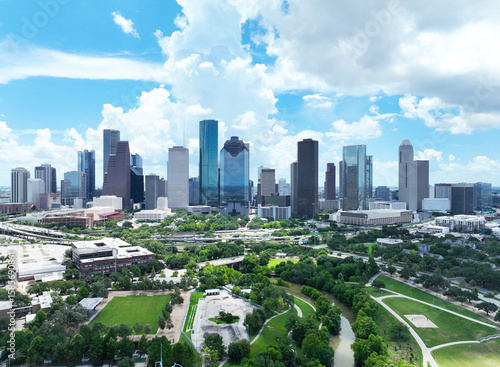 A stunning drone view of downtown Houston, Texas, showcasing the city's iconic skyline under a vibrant blue sky filled with dramatic cumulus clouds. Prominent high-rise buildings