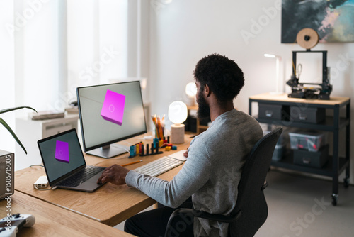 An African American man works on 3D design at his home office. He uses a laptop and monitor displaying a 3D model, with a 3D printer nearby. He is focused on his creative work.
