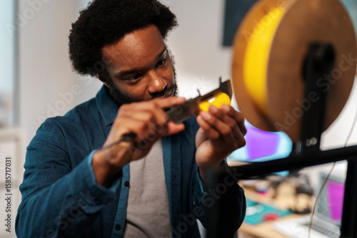 An African American man precisely measures yellow 3D printer filament with a caliper. He focuses intently on the task, ensuring accuracy for his engineering or design project.
