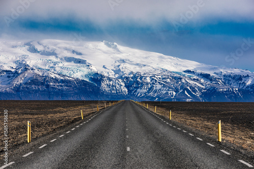 View of an endless asphalt road stretches towards a majestic snow-capped mountain range under a cloudy sky, inviting exploration and adventure, Iceland.