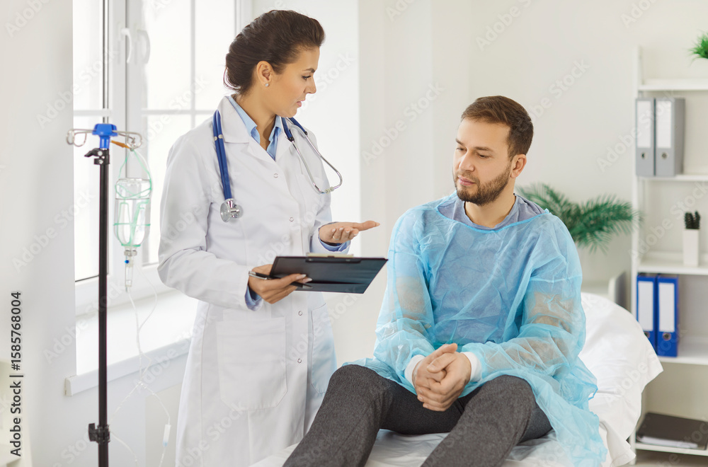 Obraz premium Female doctor in white coat discussing medical results with male patient sitting on hospital couch. Physician or cardiologist holding tablet, explaining diagnosis to young man wearing protective gown.