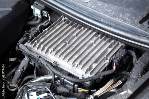 Engine control module. Close-up of an engine control unit (ECU) with ribbed silver aluminum casing. Surrounded by black insulated wires, connectors, and metal tubes in a car engine bay.