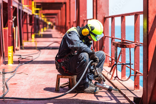 A crew member working on the deck of a merchant ship, using a pneumatic tool to remove rust.