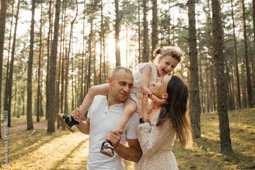 
A joyous child on father's shoulders, her mother is nearby looking up, all smiling in a sun-drenched pine forest