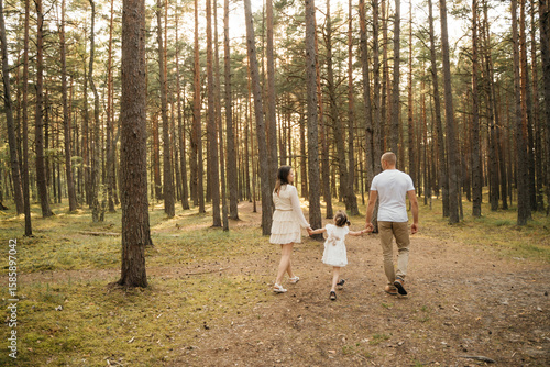 
A happy family of mother, father, and daughter walks hand-in-hand away from the camera on a sun-dappled forest path.