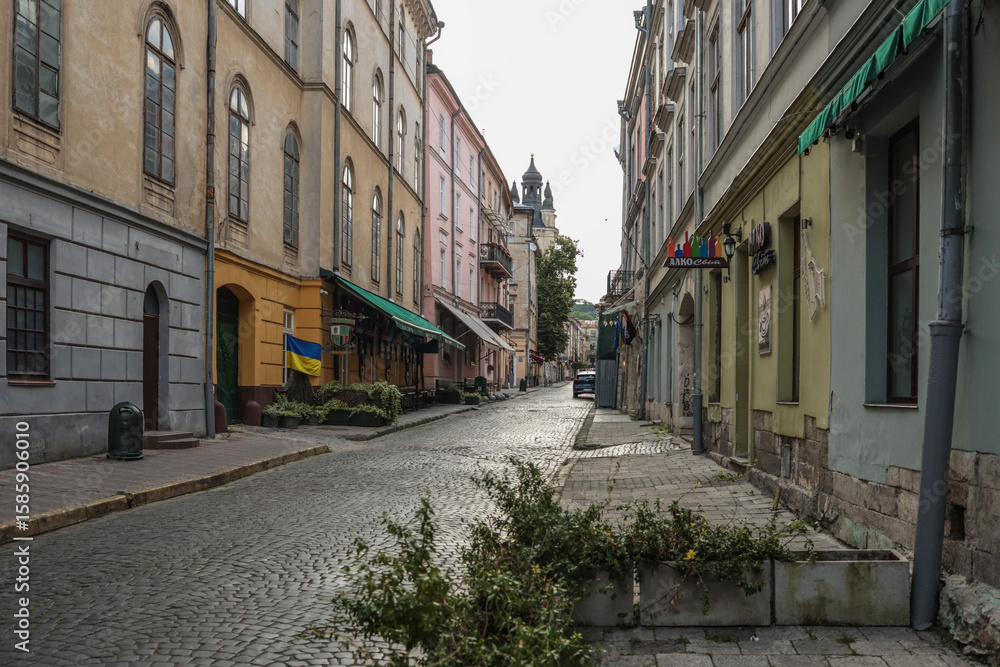 Fototapeta premium Empty cobblestone street in historic European city with colorful old buildings, Ukrainian flag, and overcast sky. Peaceful morning scene in the city center
