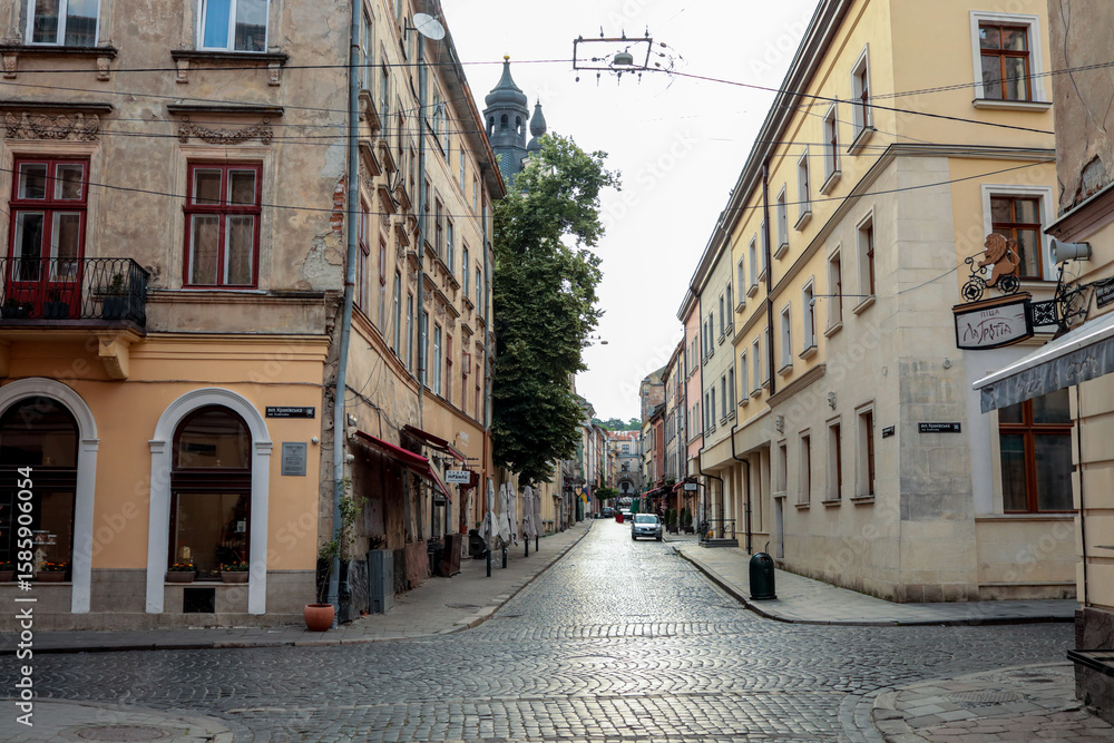 Fototapeta premium Historic European street view with cobblestone road, old buildings with arched windows and signs, and a distant church tower in the background on a quiet day