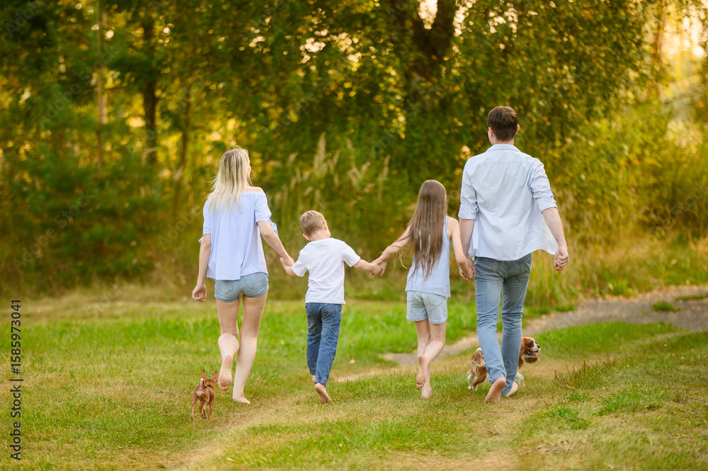 Fototapeta premium Mom, dad, boy, girl enjoying country vacation, holding hands, running barefoot on grass, jumping happily, back view. Summer family weekend concept, country vacation outside
