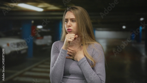 Tablou pe pânză Woman coughing in a dimly lit indoor parking lot with cars in the background, wearing a casual gray outfit, indicating distress or illness