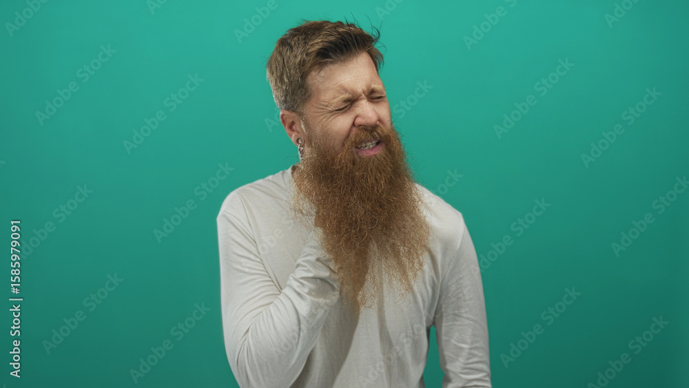 Obraz premium Man with long ginger beard in white shirt touches his throat against a green studio wall; discomfort.