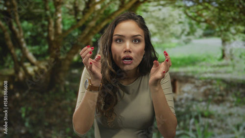 Wallpaper Mural Woman expressing surprise in a lush green park, showcasing hispanic features with red nails, seated outdoors surrounded by nature and trees, wearing a casual outfit. Torontodigital.ca