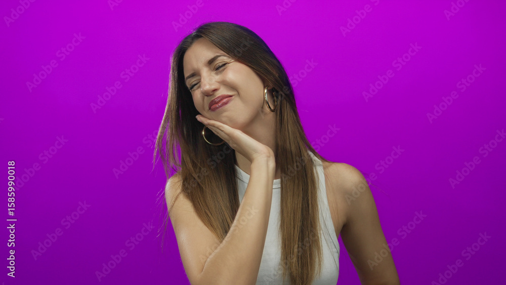 Fototapeta premium Young hispanic woman hand on cheek for toothache in purple studio setting with gold hoop earrings and white tank top; suffering health awareness prevention.