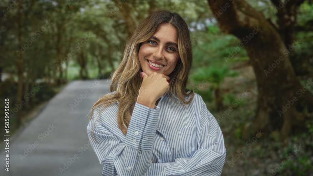Woman in striped shirt smiles while touching chin with hand on quiet forest path surrounded by trees; confidence curiosity. Woman in striped shirt smiles while touching chin with hand on quiet forest path surrounded by trees; confidence curiosity.