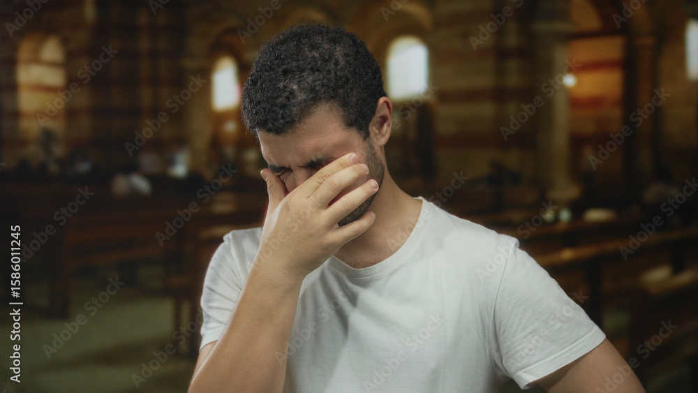 Obraz premium Man pinching nose with eyes closed in dimly lit stone church with arched windows; discomfort solitude.