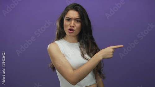 Obraz na plátně Young hispanic woman with furrowed brow pointing finger toward viewer in purple studio; rebuke anger