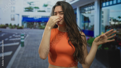 Hispanic woman wearing orange crop top pinching nose and waving hand on a crowded urban street; disgust.