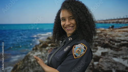 Smiling young police officer with curly hair extends hands backwards presenting sea view in studio set; pride duty.