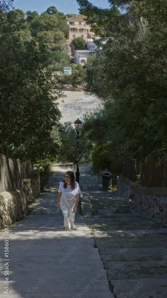 Obraz premium Woman walking up stone steps surrounded by lush greenery at beach in santanyi, mallorca with sea view in the background, capturing mediterranean essence.