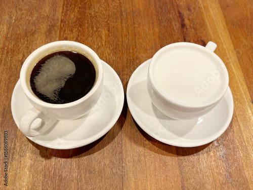 One cup of black coffee and one cup of white milk, placed side by side on a wooden table.