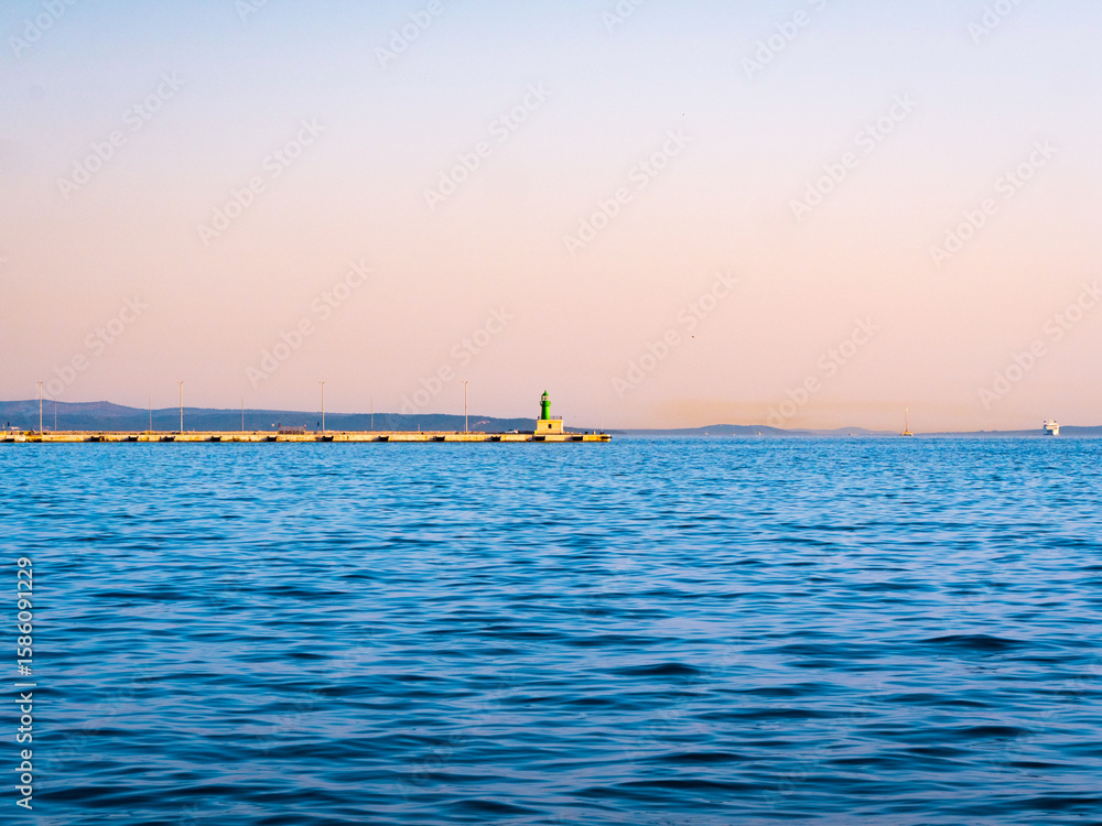 Obraz premium Split harbor in Croatia at sunset—featuring a tranquil breakwater and green lighthouse under a pastel sky, this serene coastal scene is perfect for travel and vacation inspiration.