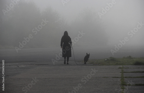 Woman walking dog in dense fog – atmospheric silhouette in moody, misty landscape