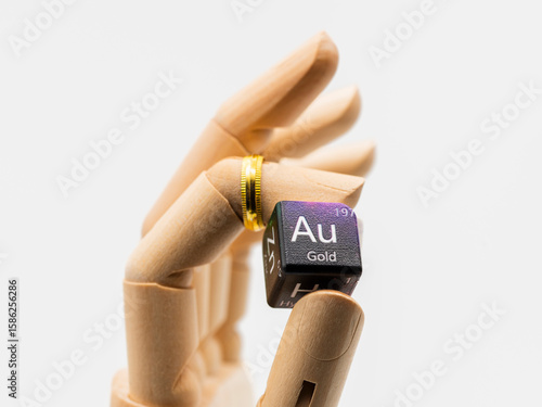 Mano de madera con un anillo de oro en el dedo sujetando un dado con el símbolo de oro de la tabla de elementos