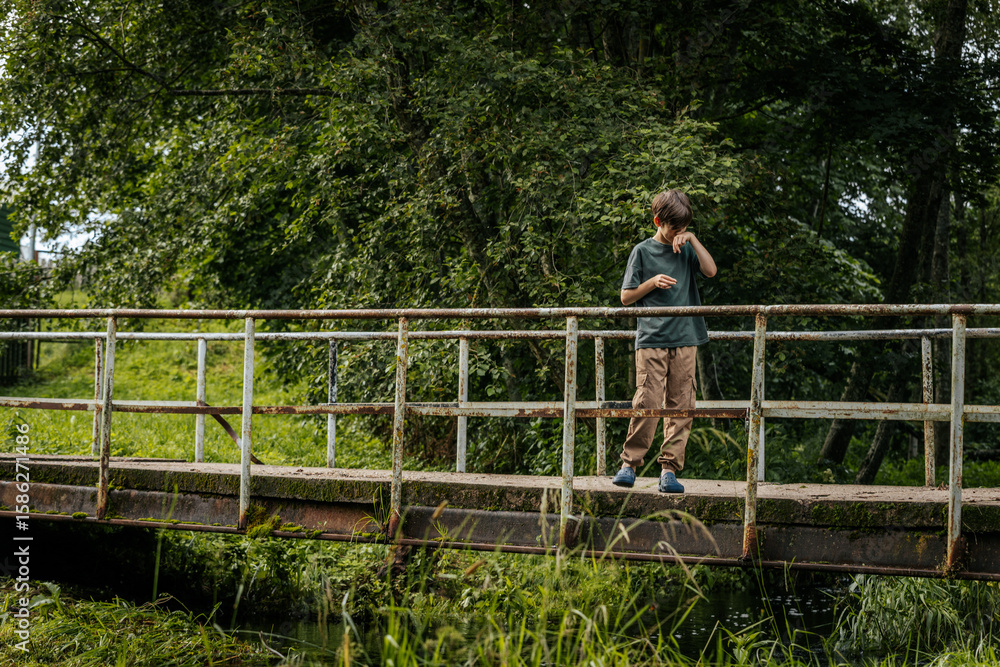 A young boy stands on an old, rusty bridge surrounded by dense foliage, wiping his face in a quiet, thoughtful moment. A young boy stands on an old, rusty bridge surrounded by dense foliage, wiping his face in a quiet, thoughtful moment.