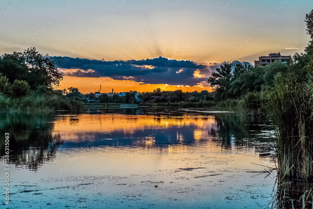 Fototapeta premium Dramatic sunset over Khorol River in Myrhorod, Poltava region, Ukraine with golden reflections on calm water surface. Peaceful riverside landscape with reeds and trees silhouetted against colorful sky