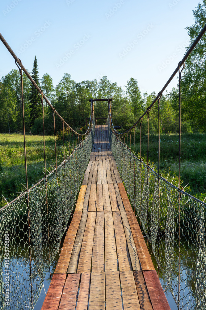 Obraz premium Suspension bridge made of wooden planks stretches across a tranquil river, surrounded by lush greenery and trees, inviting adventure and exploration in nature's serene landscape
