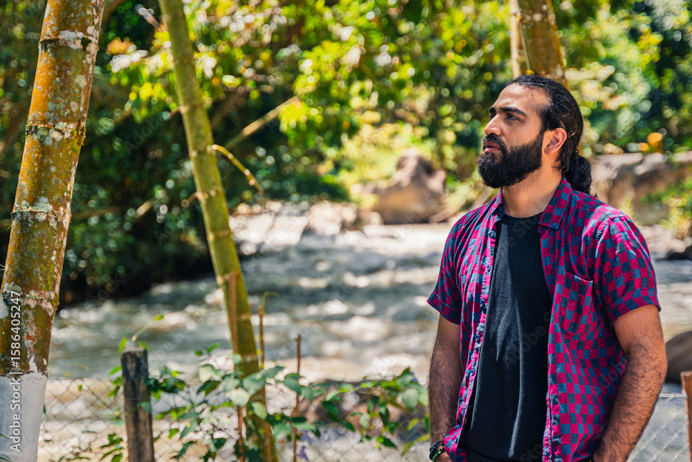 Obraz premium Contemplative Colombian man in plaid shirt standing in nature-rich forest setting near river at Santuario, Risaralda, Colombia with copy-space and rustic background