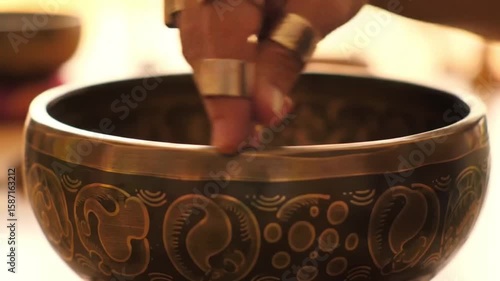 Close up of a singing bowl with golden rings on fingers resting on the edge of the bowl viewed closely