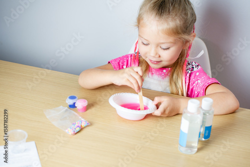Preschool girl playing with pink slime, holding wooden craft stick for mixing, focused and engaged in sensory play activity at home, DIY STEM project for kids, hands-on learning concept