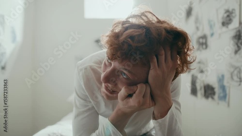 Young distressed male patient gripping his head and contorting face as experiencing psychotic episode while sitting on bed in sunlit hospital ward. Zoom-in shot