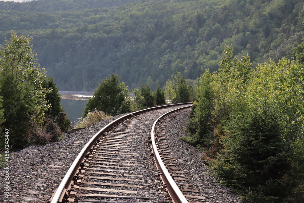 Fototapeta premium Railway in the countryside of Charlevoix in Quebec and in Canada. Summer landscape and horizon. Travel and destination and train transportation. Summer calendar with a view of a riverside.