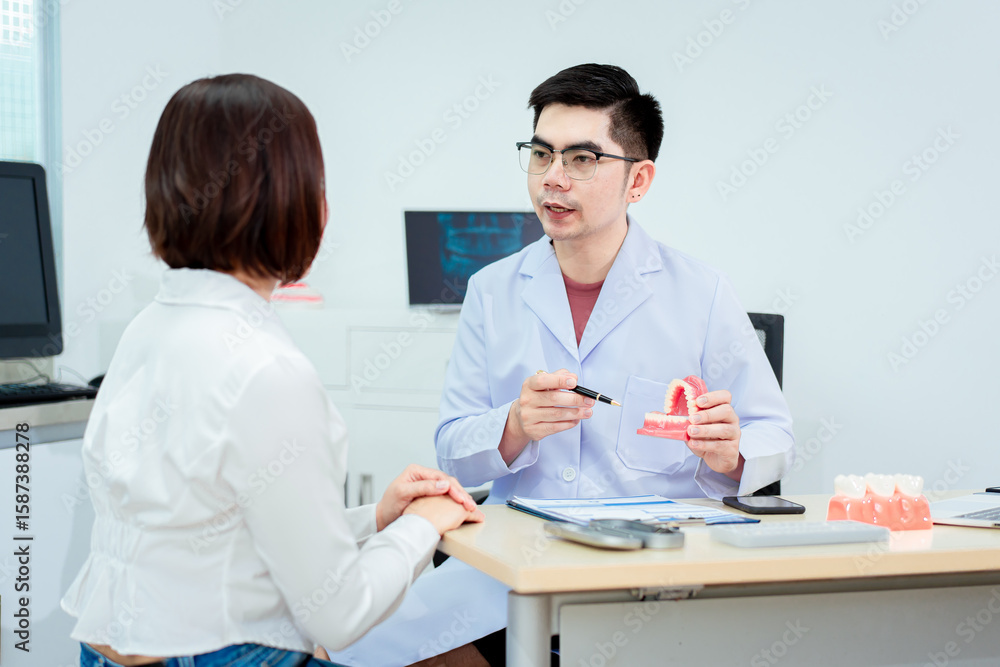Obraz premium An Asian male dentist explains dental health to a middle-aged Asian female patient while holding a dental model, demonstrating oral care and treatment procedures in a clinical consultation setting